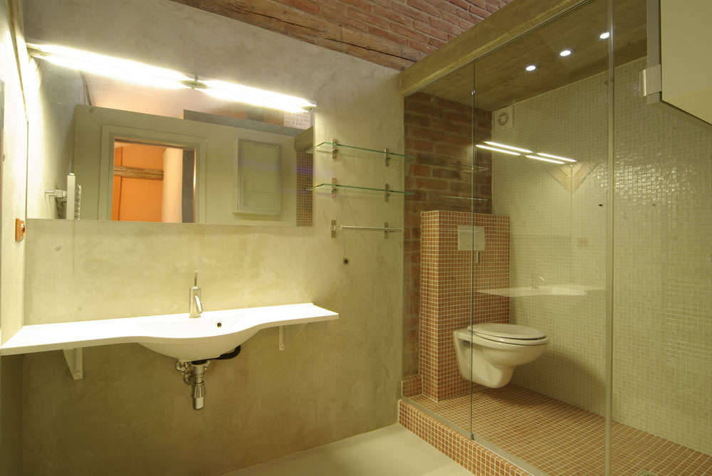 V kúpeľňovej mozaike sa akoby pripomína farebnosť a štruktúra povrchu tehlového muriva. Toaletu distingvovane oddelili od ostatnej kúpeľne sklenenou priečkou a prekryli betónovým stropom. 