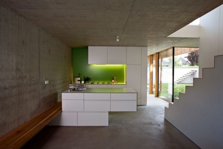 Z opačnej strany je biely kubus kuchynskou linkou. Vďaka vstavanému nábytku sa obytné priestory vyčistili a pôsobia voľne a veľkoryso, farebná kombinácia bielej, sviežej zelenej a dreva, typická pre celý dom, vnáša do interiéru optimistickú atmosféru.