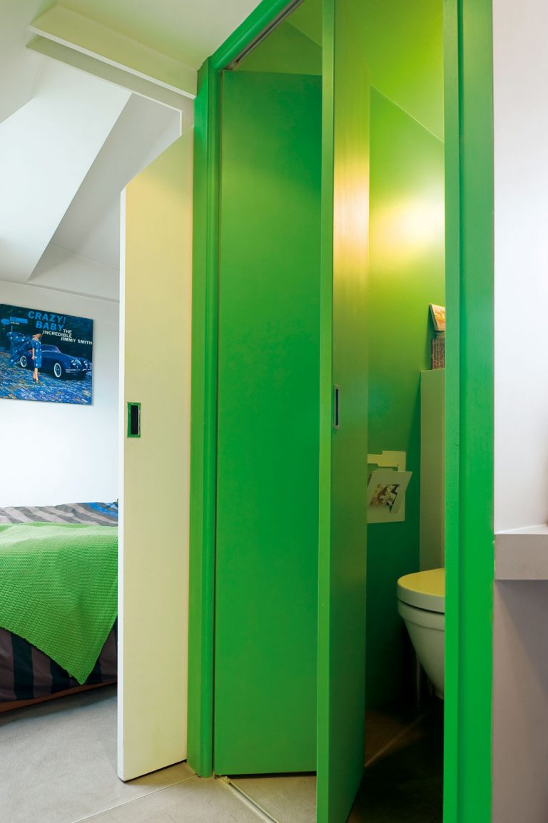 Hneď vedľa spálne je za zelenými skladacími dverami toaleta. Rovnako ako posuvné dvere spálne, prispôsobené sklonu strechy, sú aj dvere WC vyrobené na mieru. Aj v tomto prípade myslel architekt na to, ako čo najpraktickejšie využiť daný priestor. (Pletená prikrývka na posteli je od Zara Home.)