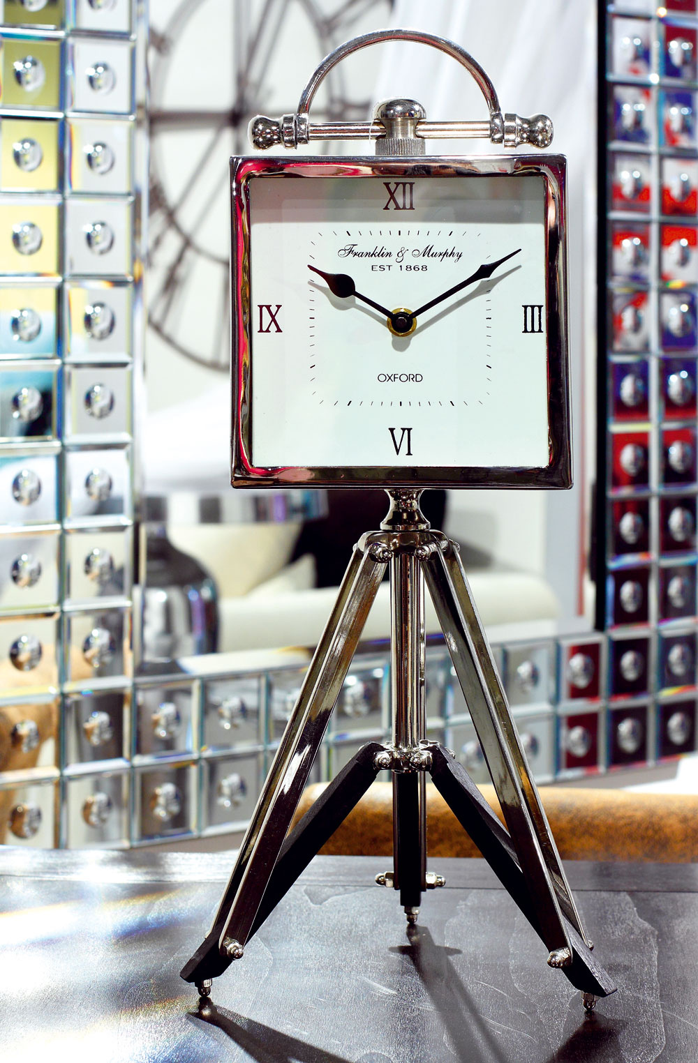 Stolové hodiny Oxford Square na stojane s pohyblivou výškou. Cena 135,90 €. V pozadí nástenné hodiny Clock Factory s rímskymi číslicami, vyrobené z tvárneho železa natretého na hnedo. Priemer 120,2 cm. Cena 118,90 €. Predáva Kare, LightPark.