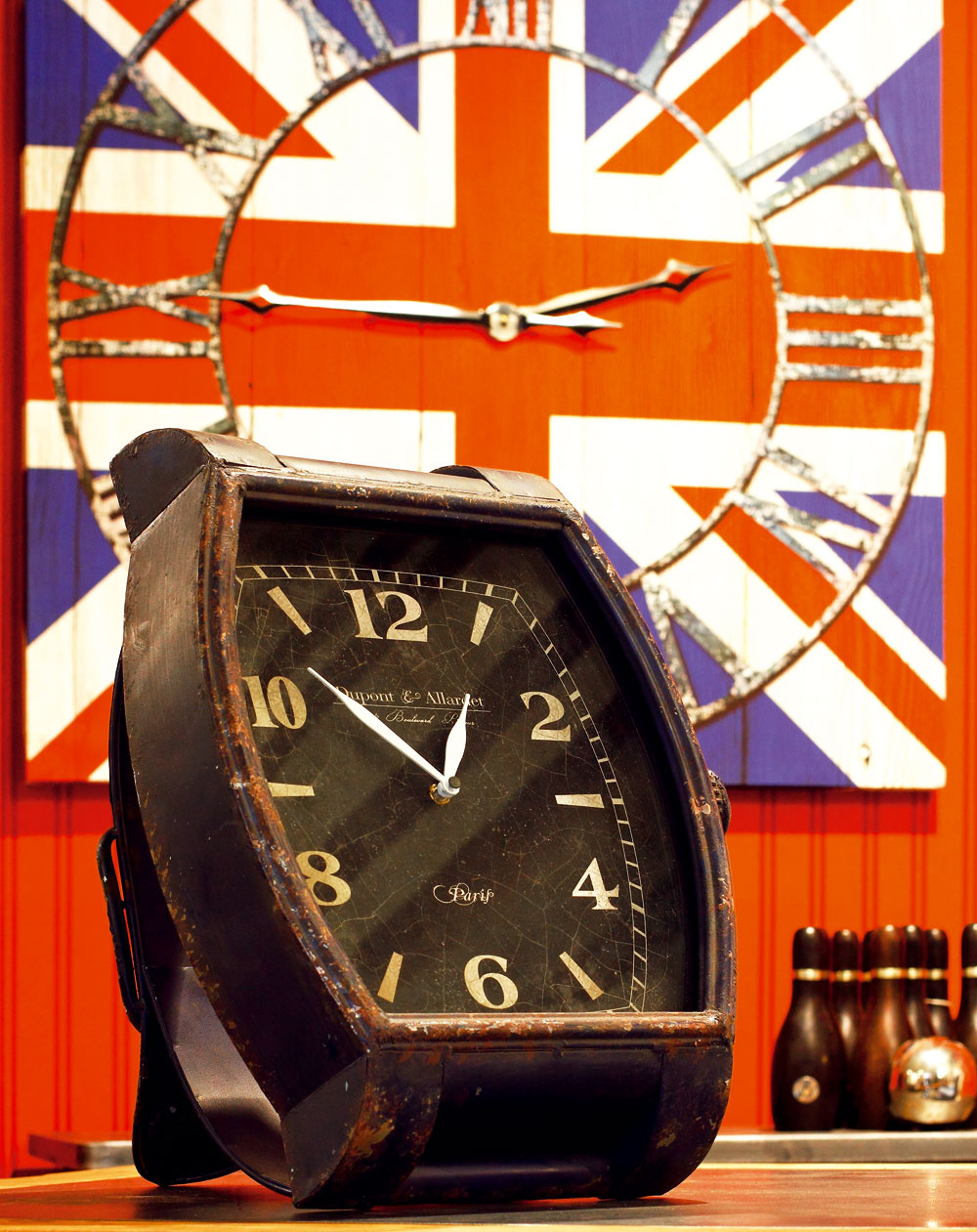 Stolové „náramkové“ hodiny Vintage Blue z modrého kovu so zaskleným ciferníkom. Rozmery: 38,5 × 34,5 × 33,5 cm. Cena 109 €. V pozadí hodiny – obraz Very British s motívom britskej vlajky. Rozmery 90 × 90 cm. Cena 75,90 €. Predáva Kare, LightPark.