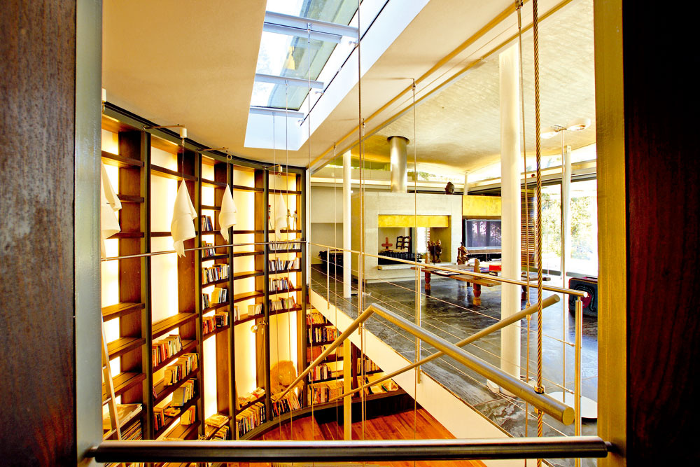 Pohľad na schodište do knižnice z chodby, ktorá vedie ku krytým schodom do horných pavilónov. Okno, ktoré schody osvetľuje, vyúsťuje v trávniku pred spálňami na najvyššom podlaží. S oceľovým zábradlím ladia perforované stupne zavesené na lanách. Stena knižnice je jedným z mnohých oblúkov v dome a okolo neho. Na konci obývačky žiaria odrazeným svetlom mosadzné panely – oživujú stenu s kozubom v betónovom rámci.