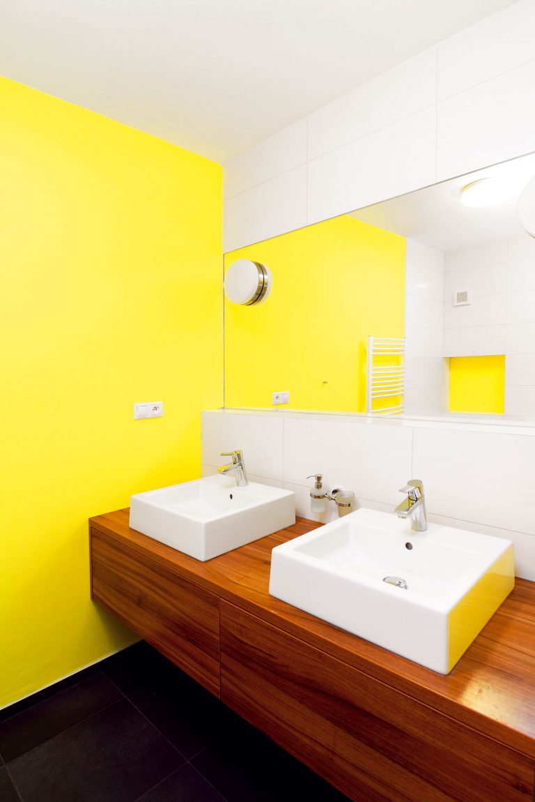 Nová kúpeľňa vznikla na rovnakom pôdoryse ako jej predchodkyňa. Overená kombinácia farieb: čierna podlaha, biele obklady, žlté steny a drevo, pôsobí útulne a čisto zároveň a priestoru bez prirodzeného osvetlenia dodáva teplo aj pozitívnu energiu.
Nika pri vani je určená na odloženie hygienických potrieb, ktoré treba mať poruke, v zásuvke umývadlovej skrinky je ďalší odkladací priestor na väčšie a menej často používané predmety, napríklad čistiace prostriedky. Keďže miestnosť nemá prirodzené vetranie, uloženie uterákov a bielizne tu architekti neodporúčali.