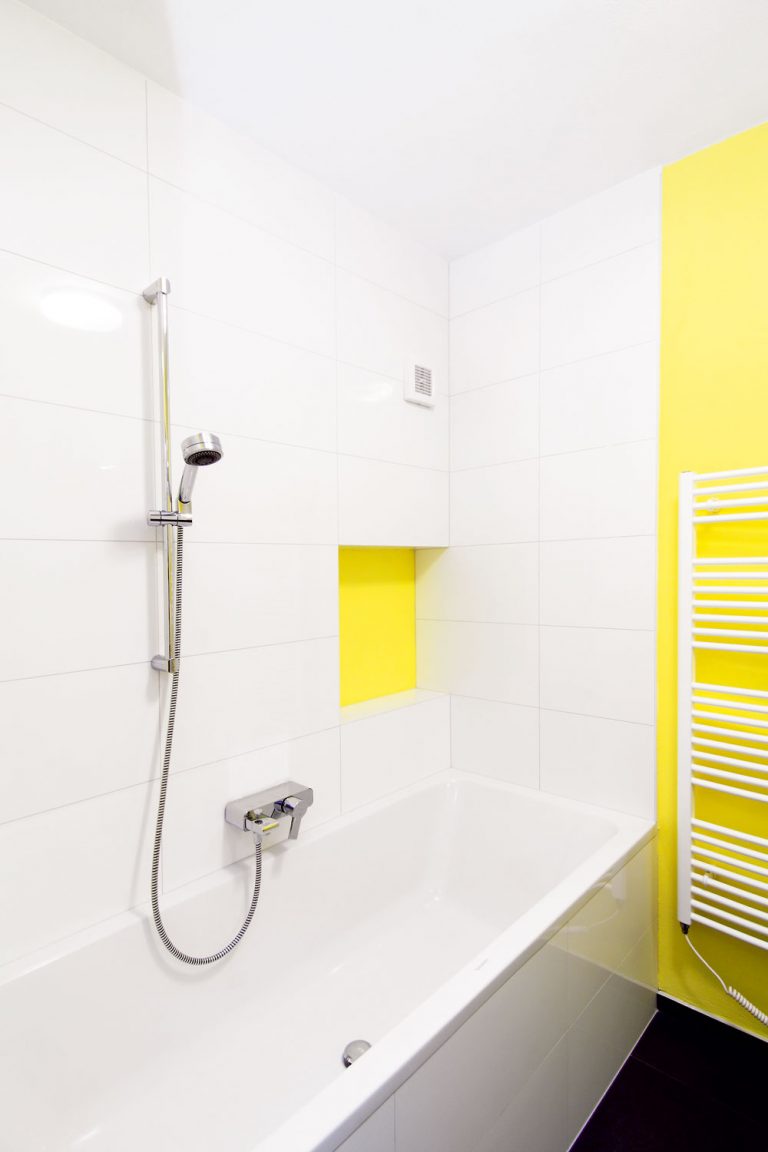Nová kúpeľňa vznikla na rovnakom pôdoryse ako jej predchodkyňa. Overená kombinácia farieb: čierna podlaha, biele obklady, žlté steny a drevo, pôsobí útulne a čisto zároveň a priestoru bez prirodzeného osvetlenia dodáva teplo aj pozitívnu energiu.
Nika pri vani je určená na odloženie hygienických potrieb, ktoré treba mať poruke, v zásuvke umývadlovej skrinky je ďalší odkladací priestor na väčšie a menej často používané predmety, napríklad čistiace prostriedky. Keďže miestnosť nemá prirodzené vetranie, uloženie uterákov a bielizne tu architekti neodporúčali.