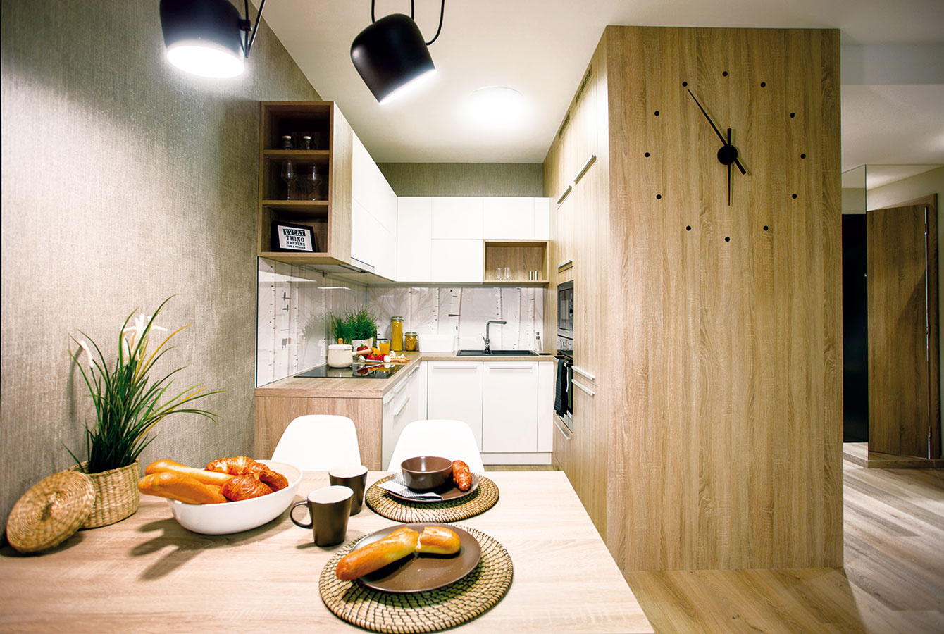 Premyslené stretnutie. Kuchyňa menšieho z bytov je praktická „biela klasika“, ktorú architekti skombinovali s drevenými prvkami. Smerom do obývačky je linka ukončená drevenými poličkami, kde môžu byť knižky, doplnky, obrázky – táto časť tak vyzerá menej kuchynsky a viac komunikuje s obývačkou. 