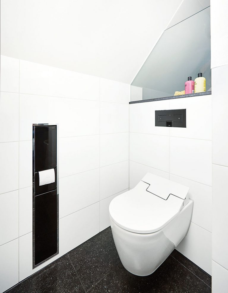 Investor si želal, aby sa aj zariadenie kúpeľne prispôsobilo bielo-čiernej farebnej kombinácii, ktorá je príznačná pre celý interiér rodinného domu. Tá sa objavuje aj v priestore toalety, kde sa nachádza čierne splachovacie WC tlačidlo vsadené do bieleho obkladu.