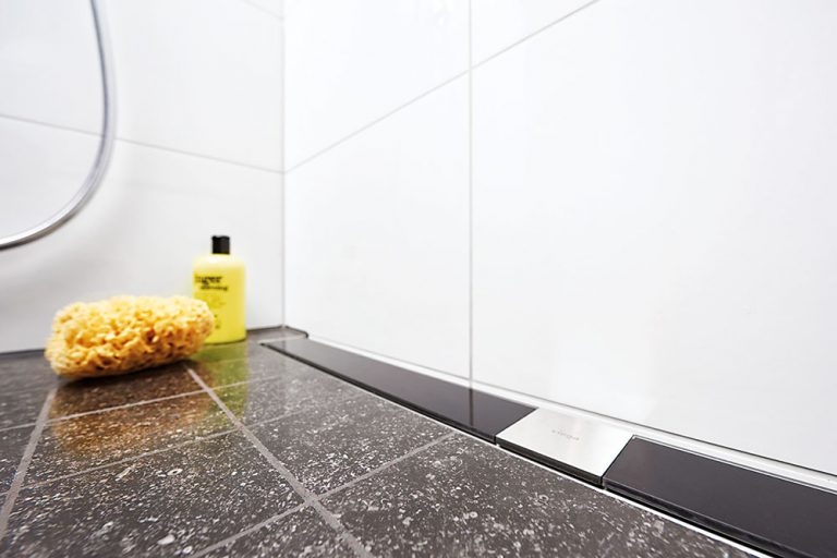Sprchový žľab je harmonicky začlenený do podlahy. Projektant v tomto prípade zvolil odtok k stene a rám z nehrdzavejúcej ocele s dizajnovým roštom z čierneho bezpečnostného skla.