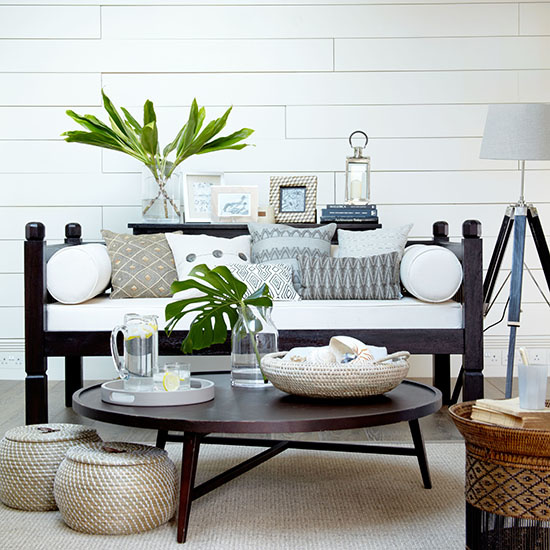 All white living room, dark wood sofa bench with white cushion, selection of cushions, large round dark wood coffee table, wicker baskets, neutral rug, banana leaves in vase, white panelled walls, tripod floor lamp. IH 08/2011 pub orig