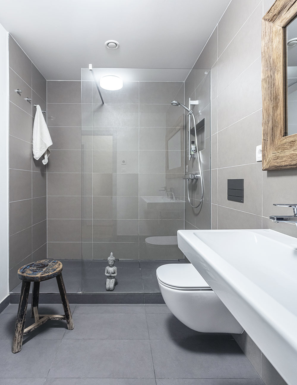 Kúpeľňa je obložená sivým obkladom a so zvyškom bytu ju štýlovo spájajú drevené prvky, ako sú rám zrkadla nad umývadlom či praktická stolička.