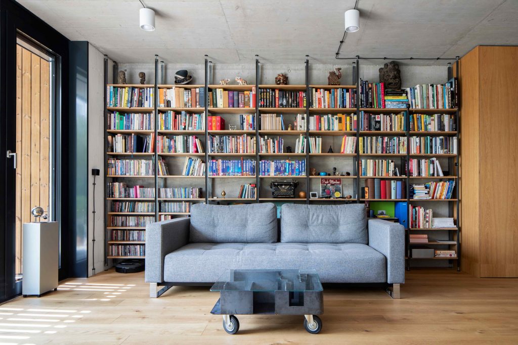 Obývačka s veľkou knižnicou sivým gaučom a betónovým stropom