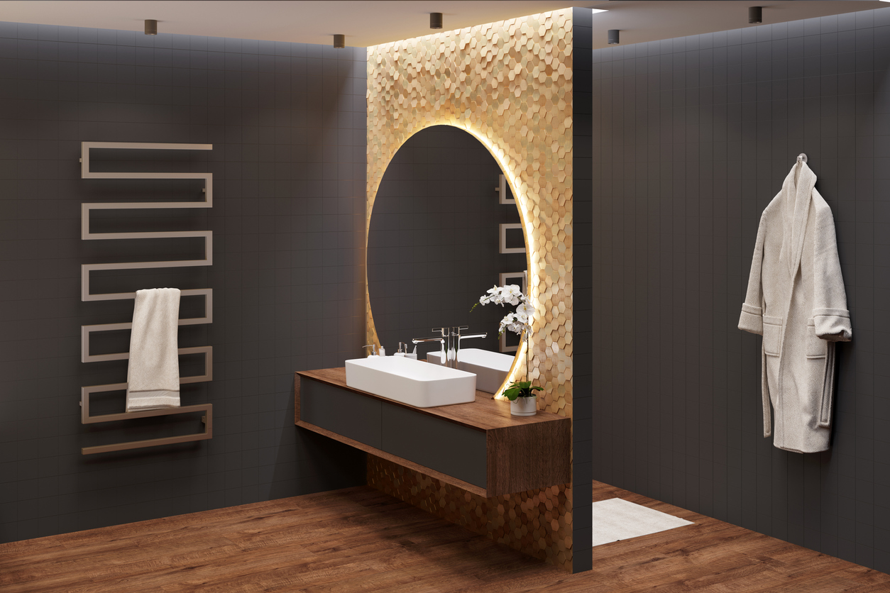 Bathroom with black walls and wooden floor, with a washbasin next to a large round mirror on the wall with gold mosaic, a towel on a heated towel rail, a terry robe on a black wall.