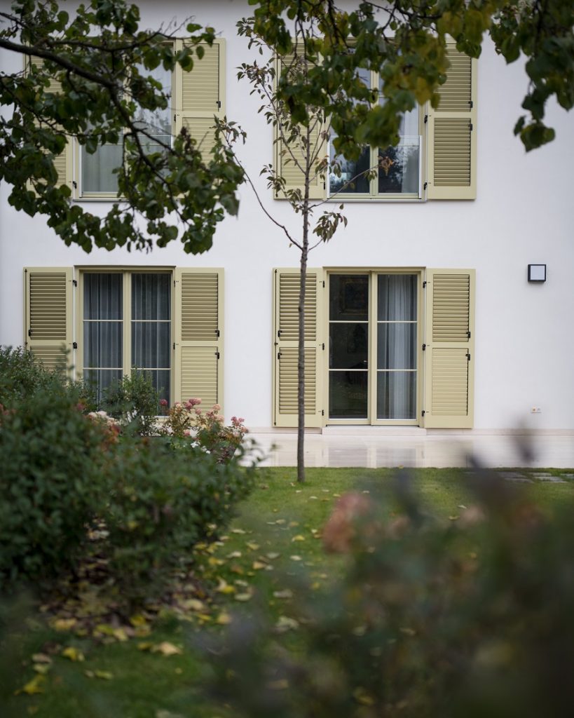 dom s bledožltými krídlovými okenicami, pohľad spredu zo záhrady