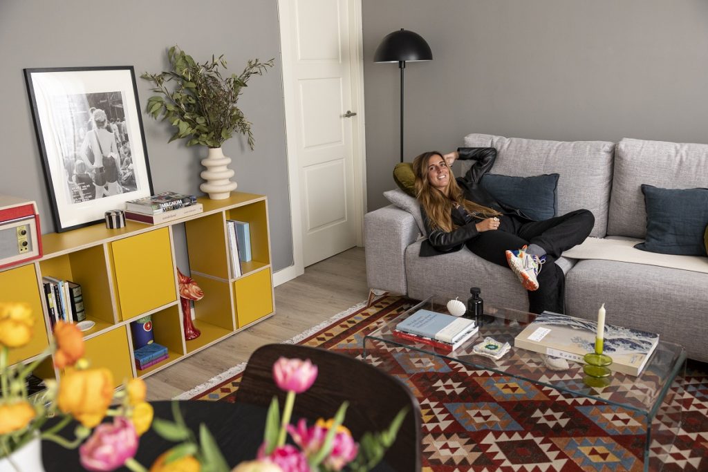 Žena Elisa Serrano leží na sivej pohovke v obývačke s pestrým kobercom a žltou komodou.