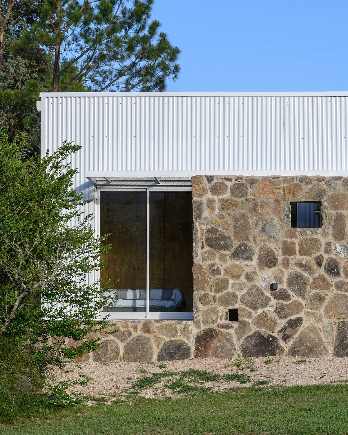 Kamenný nie je len vstupný múr. Celý dom stojí na podstavci z kameňa, čím architekti dosiahli silnejší súlad s krajinou. Samotné telo domu je obalené súvislým plechovým plášťom v bielej farbe. 