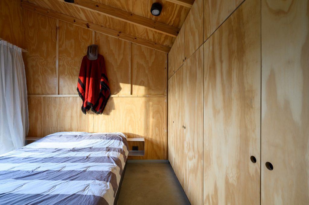 Spálňa s posteľou v drevostavbe, na vešiaku klobúk a červený odev