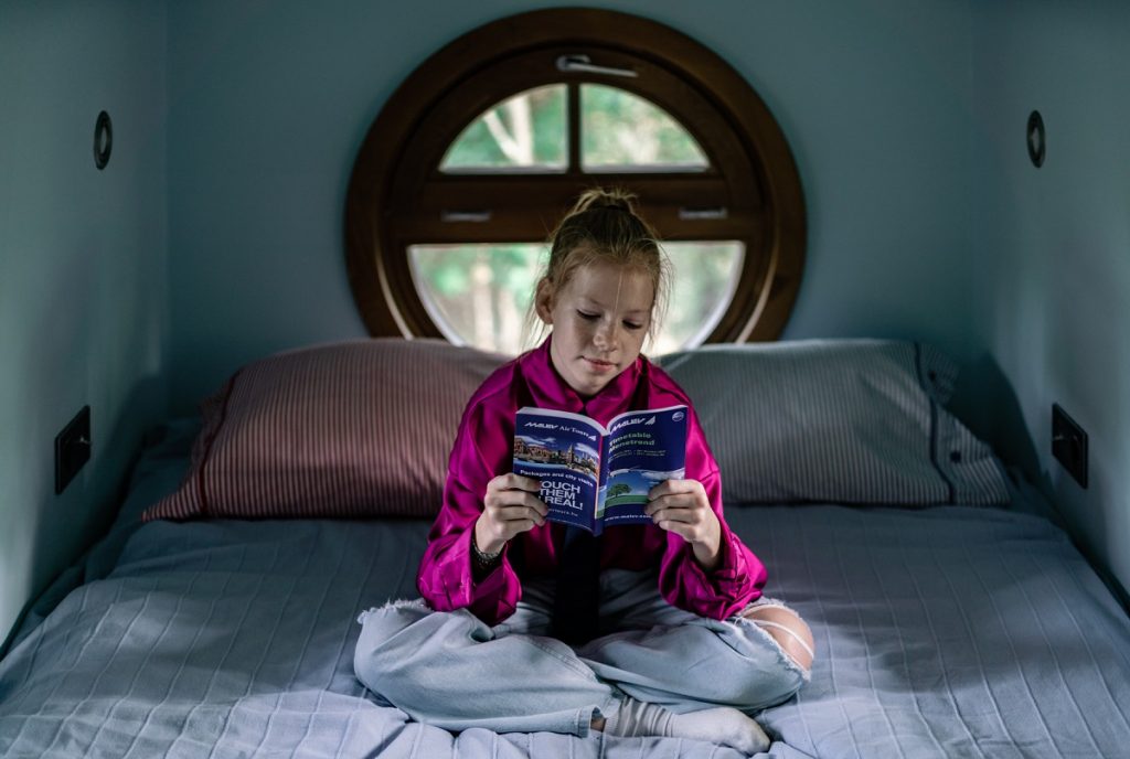 dievčatko sedí na posteli s knižkou, vzadu okrúhle okno