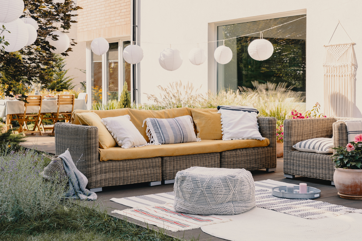 Pouf next to rattan couch and armchair on wooden terrace with flowers and lamps