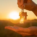 The hand holds the key to the new house with a keychain in the form of a house on the background of the sunset