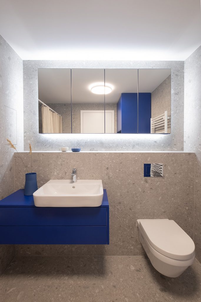 Modrá kúpeľňová skrinka so sivým veľkoformátovým obkladom na stenách