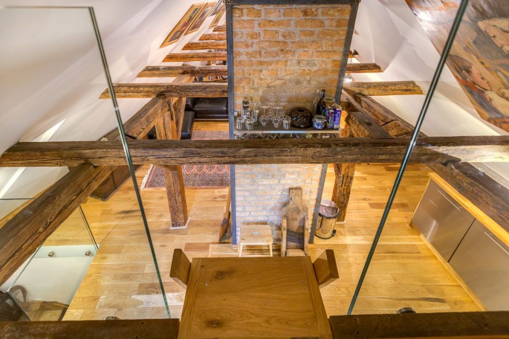 Pohľad zvrchu dreveného rebríka na priestor o poschodie nižšie cez viditeľné drevené trámy.