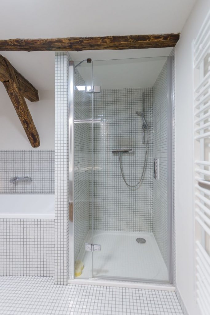 Biela kúpeľňa s bielou mozaikou na stenách i podlahe, na strope viditeľné drevo. Pohľad na sprchovací kút.