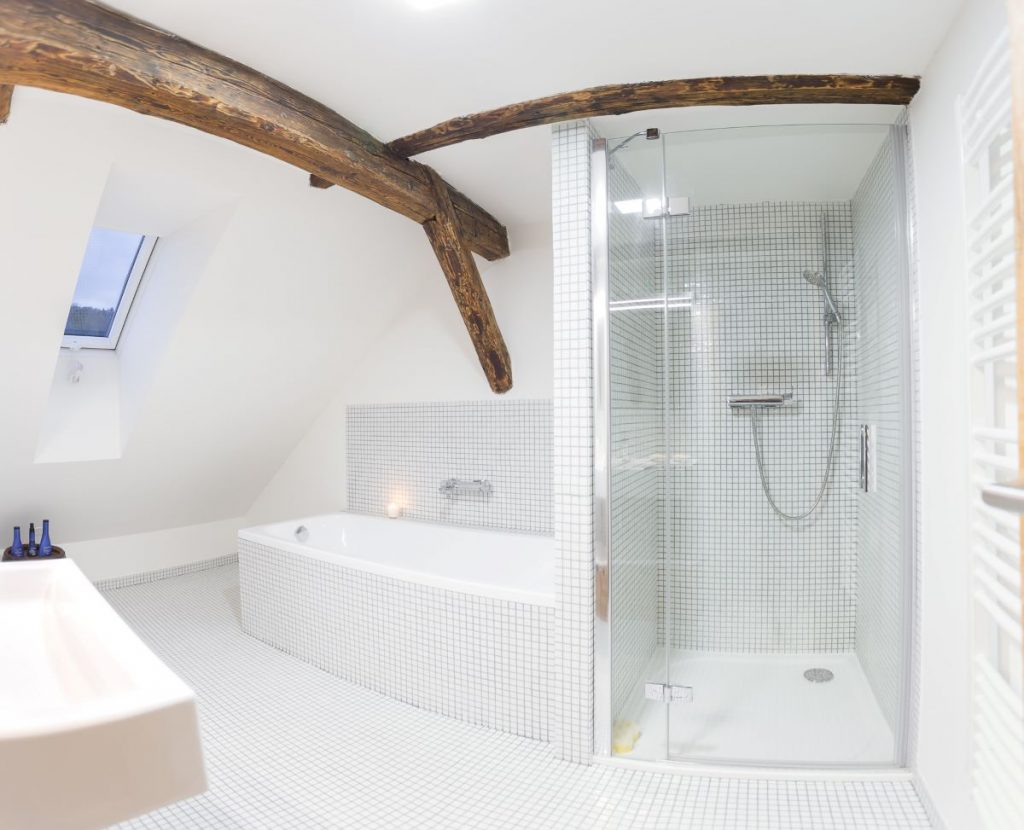 Biela kúpeľňa s bielou mozaikou na stenách i podlahe, na strope viditeľné drevo. Pohľad na sprchovací kút a vaňu.