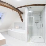 Biela kúpeľňa s bielou mozaikou na stenách i podlahe, na strope viditeľné drevo. Pohľad na sprchovací kút a vaňu.