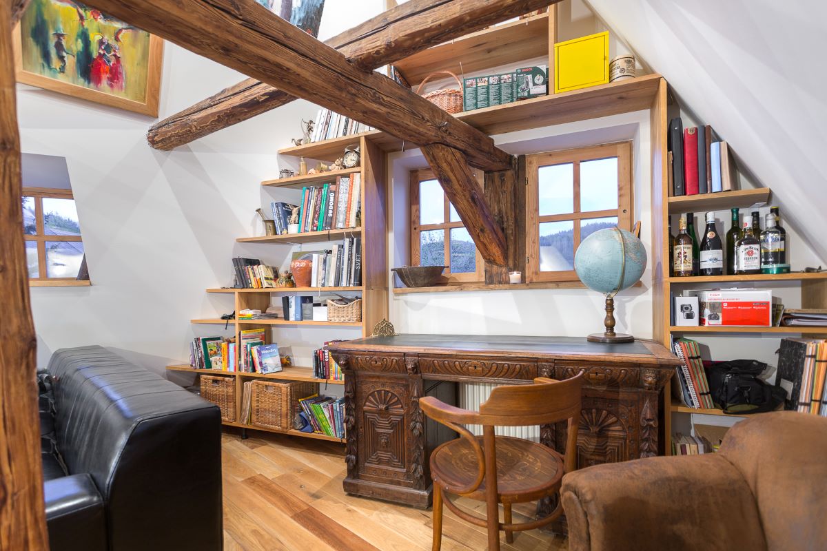 Na mieru robená knižnica obkolesujúca malé štvorcové okná, pod ňou historický drevený stôl a drevená stolička.