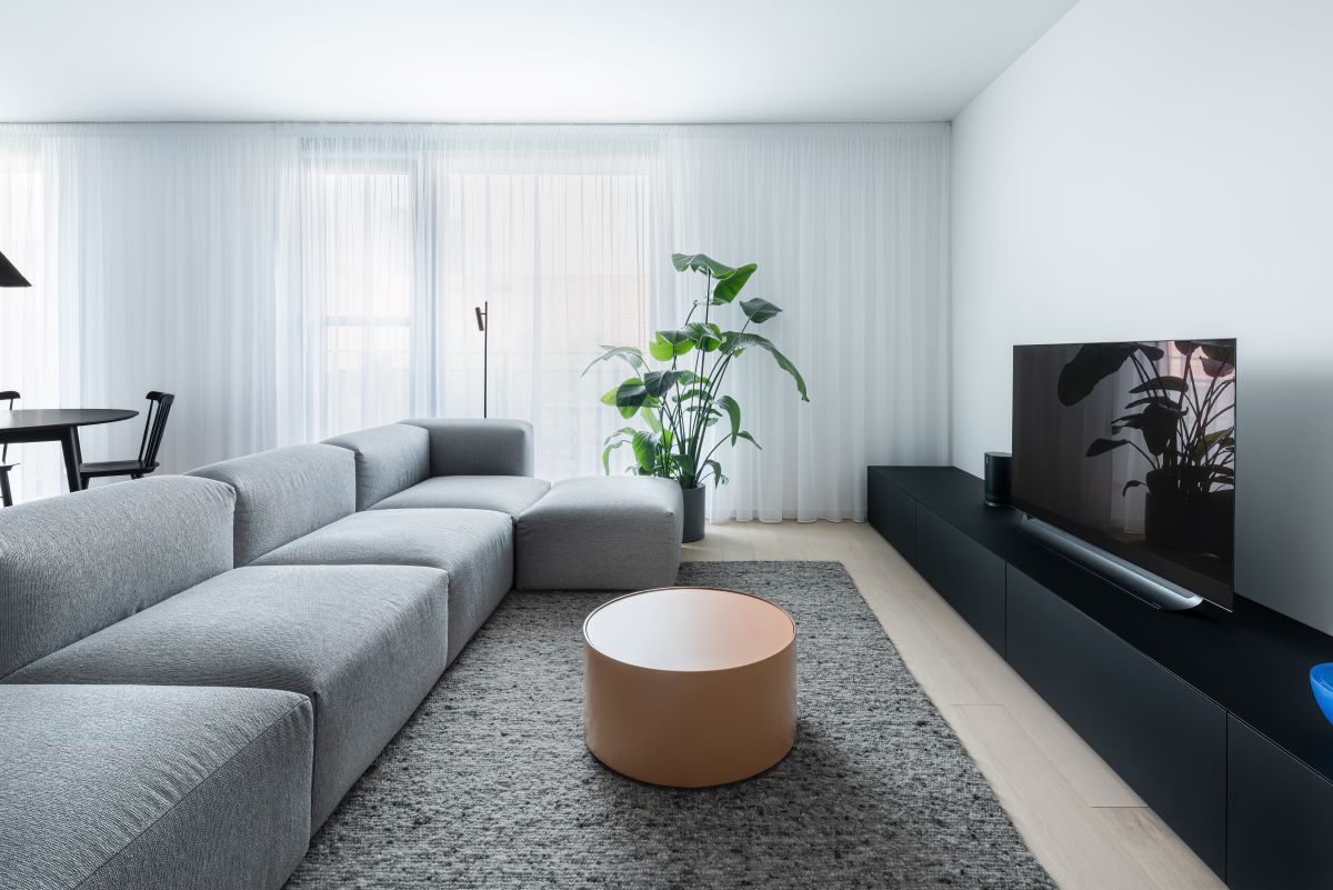 Obývačka v byte so svetlosivou sedačkou, sivým kobercom, čiernou televíznou skrinkou a okrúhlym konferenčným stolíkom v lososovej farbe.