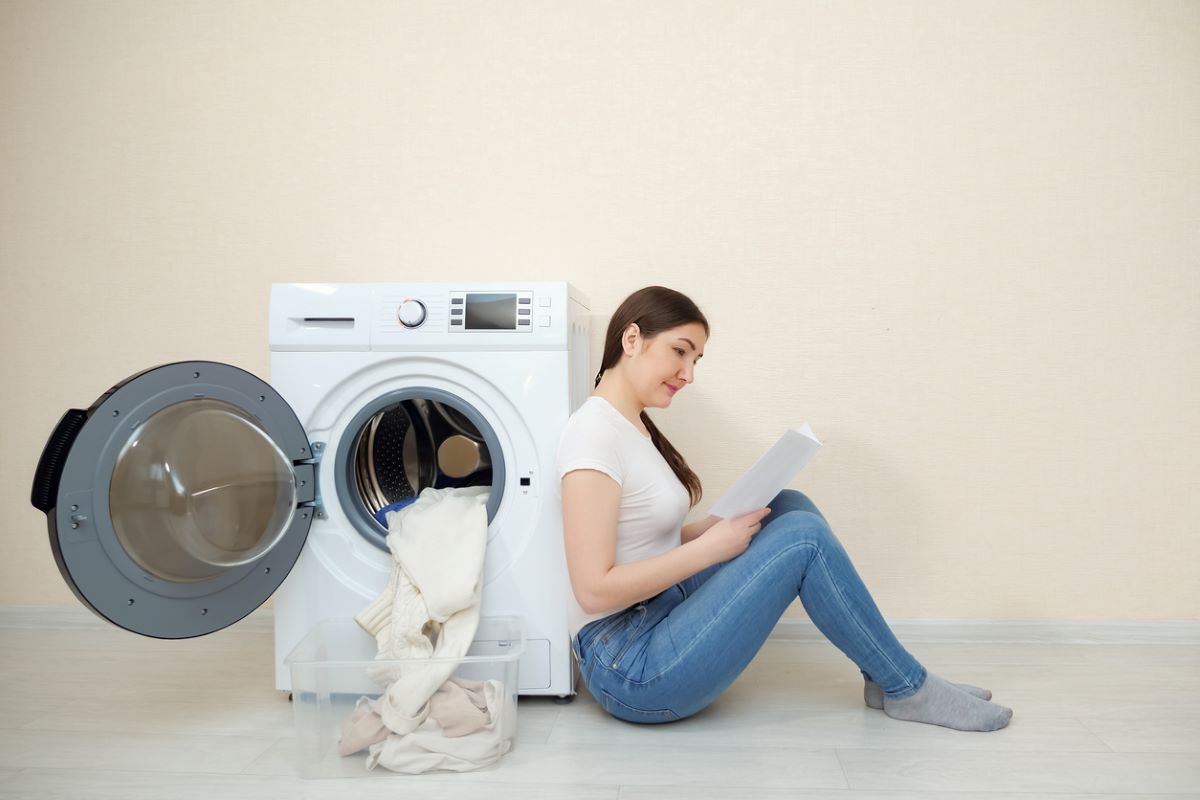 Žena sedí vedľa práčky s otvorenými dvierkami, z ktorých vypadáva bielizeň, a číta návod na použitie.