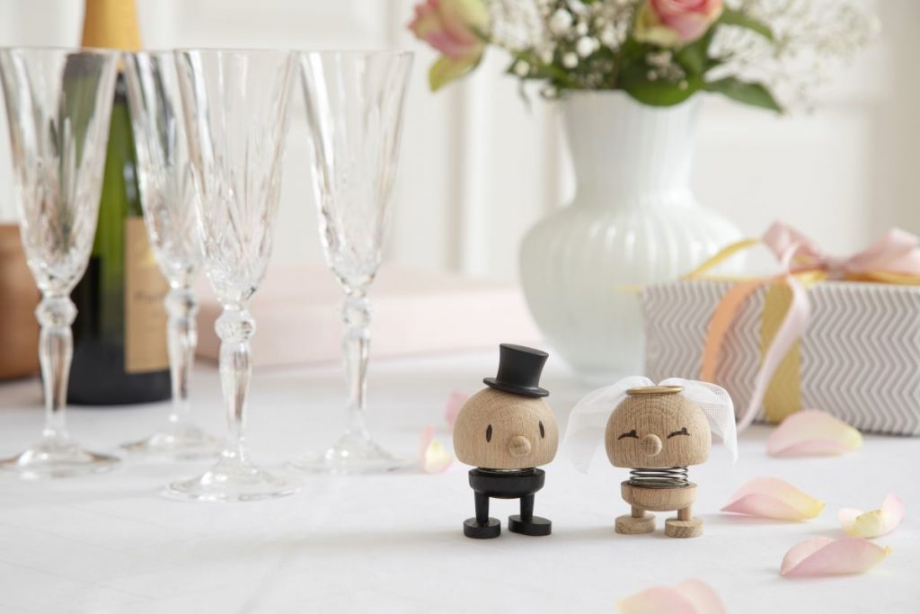 Drevené figúrky hoptimisti Bride nad Groom od Cross Fingers na stole s biely obrusom, v pozadí biela váza s kvetmi a poháre na stopke.