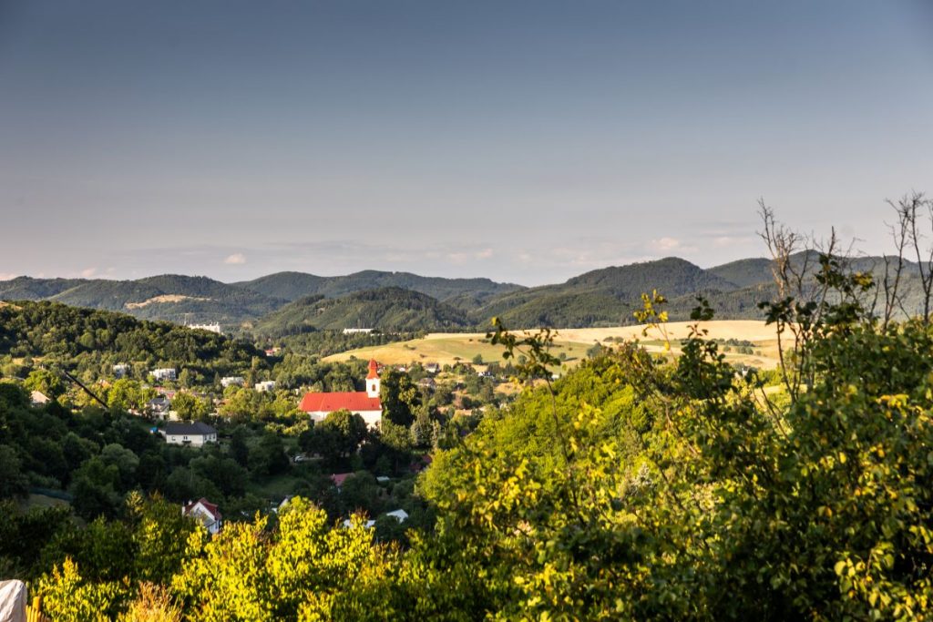 Výhľad na okolie Banskej Štiavnice s domami ukrytými medzi stromami a vystupujúcim kostolom s červenou strechou, v pozadí Nízke Tatry.