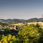 Výhľad na okolie Banskej Štiavnice s domami ukrytými medzi stromami a vystupujúcim kostolom s červenou strechou, v pozadí Nízke Tatry.