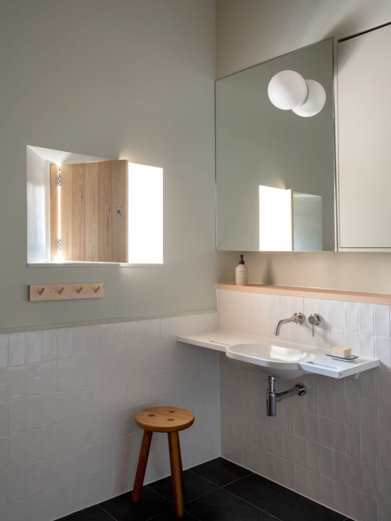 Kúpeľňa v interiéri s obkladom z bielych kachličiek v dolnej tretine stien, bielym umývadlom, zrkadlom a drevenou trojnožkou.