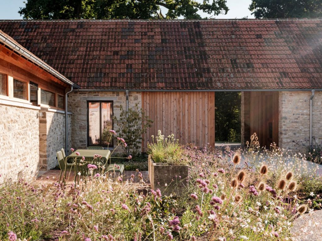 Exteriér farmárskej budovy so zeleným záhradným nábytkom a bujným záhonom.