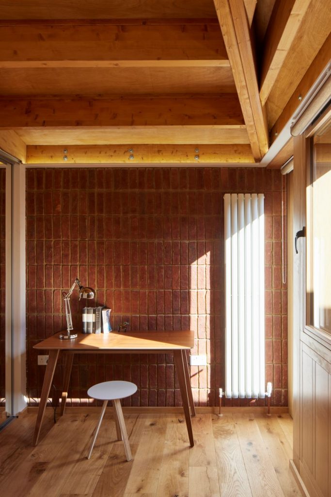 Drevený písací stôl s nízkou okrúhlou stoličkou pri tehlovej stene v interiéri bungalovu.