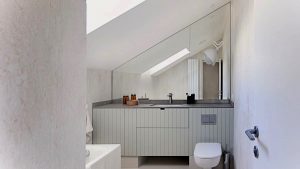 Kúpeľňa od Linea Design s veľkým lichobežníkovým zrkadlom, toaletou a skrinkou s umývadlom.