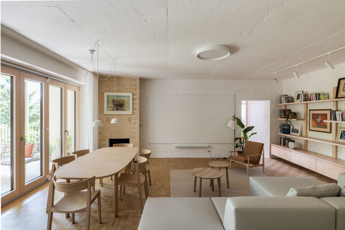 Obývačka s premietacou stenou, oválnym dreveným stolom so stoličkami, kozubom so šamotovým obkladom, svetlou sedačkou a policami na stene.
