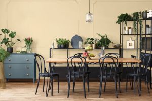 Jedálenský stôl s drevenou doskou a čiernou konštrukciou, pri ktorom je šesť čiernych stoličiek. V pozadí pri stene tmavá modrošedá komoda a čierna polica plná rastlín
