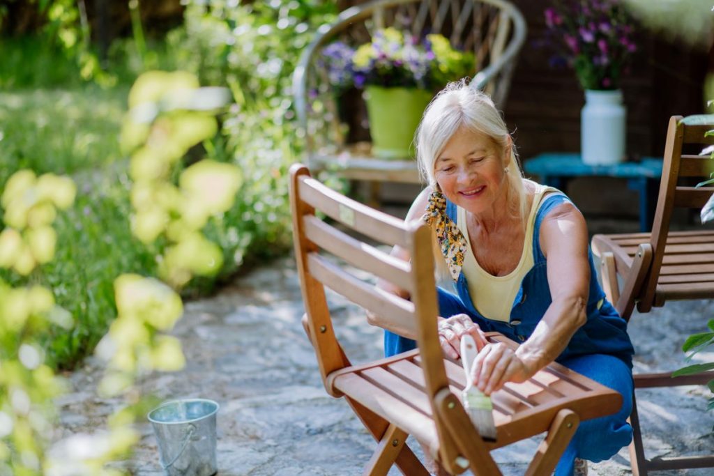 Staršia žena v žltom tielku a nohaviciach na traky lakuje drevenú záhradnú stoličku v exteriéri.