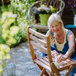 Staršia žena v žltom tielku a nohaviciach na traky lakuje drevenú záhradnú stoličku v exteriéri.