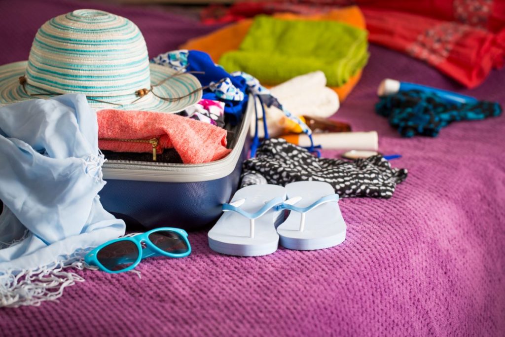 Časť otvoreného kufra s obsahom na letnú dovolenku, ako klobúk, slnečné okuliare či flip-flopy na fialovej deke.