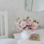 Kytica svetloružových kvetov v bielej váze z džbánu na bielej komode. Na svetlej stene s kvetinovým vzorom je zavesené zrkadlo.