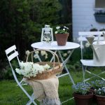 Biely záhradný nábytok pred záhradným domčekom s pleteným košíkom s kvetmi, petrolejkou a kvetmi v kvetináči.