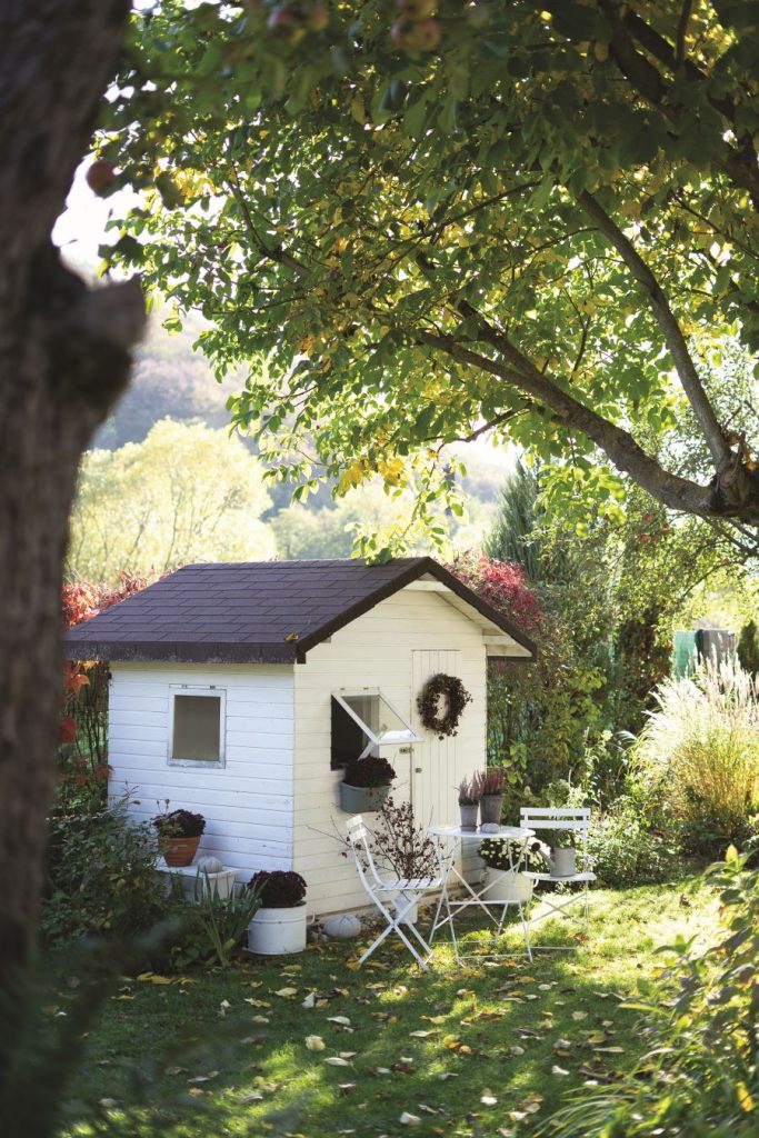 Drevený záhradný domček s tmavou strechu a subtílnym záhradným nábytkom pred dverami.