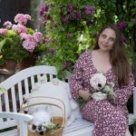 Alexandra v kvetovaných šatách s kvetmi v ruke sedí na bielej drevenej lavičke medzi kvetinovým záhonom, vedľa nej sú biele okrúhle podušky so strapcami a pletený košík s bielym džbánom.