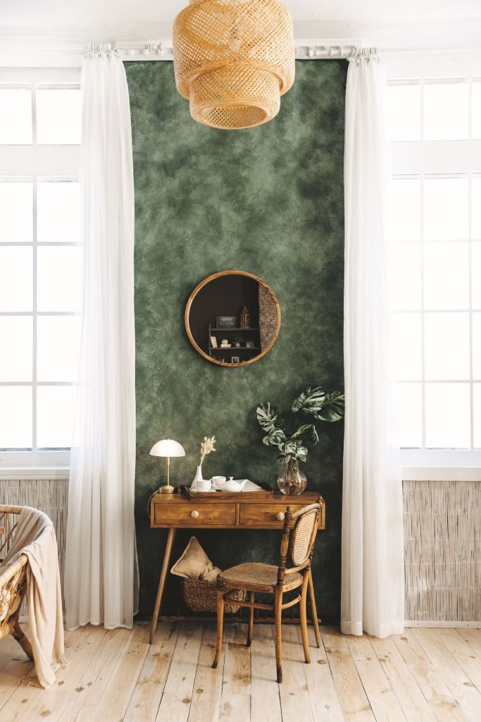 Drevený stolík so zásuvkami a drevenou stoličkou pri tmavozelenej stene s okrúhlym zrkadlom.