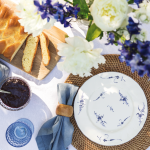 Stôl s bielym obrusom, bielym tanierom s modrým kvetovaným vzorom, modrou utierkou, prírodným prestieraním a kvetmi. Naservírovaná je vianočka s džemom.
