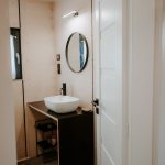Kúpeľňa v drevenom domčeku s umývadlom položeným na čiernej doske a okrúhlym zrkadlom nad ním.