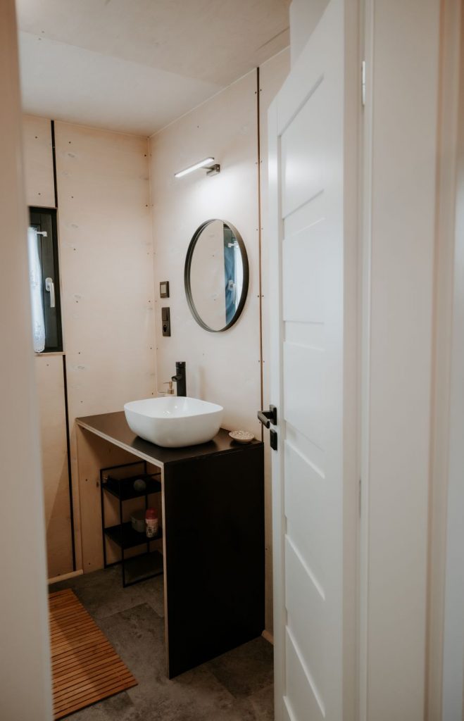 Kúpeľňa v drevenom domčeku s umývadlom položeným na čiernej doske a okrúhlym zrkadlom nad ním.